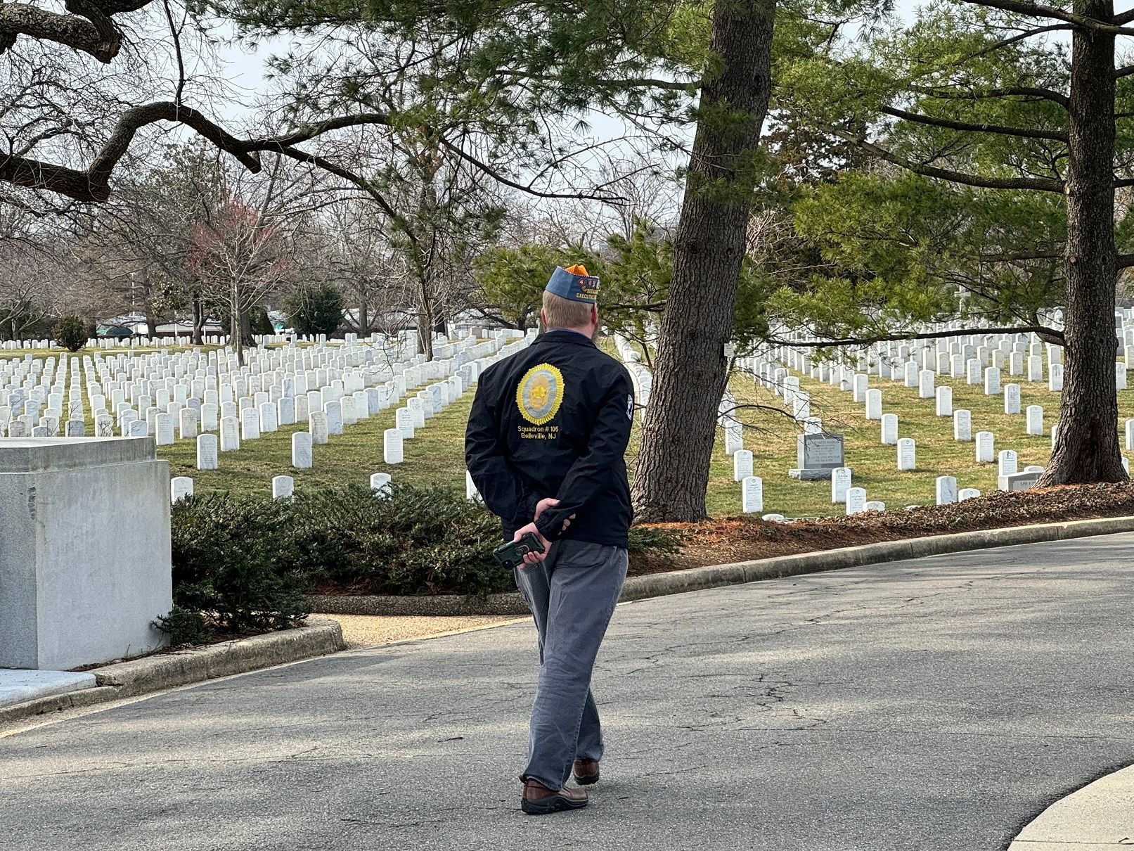 Man in uniform jacket walks through a cemetery with white headstones; sunny day.