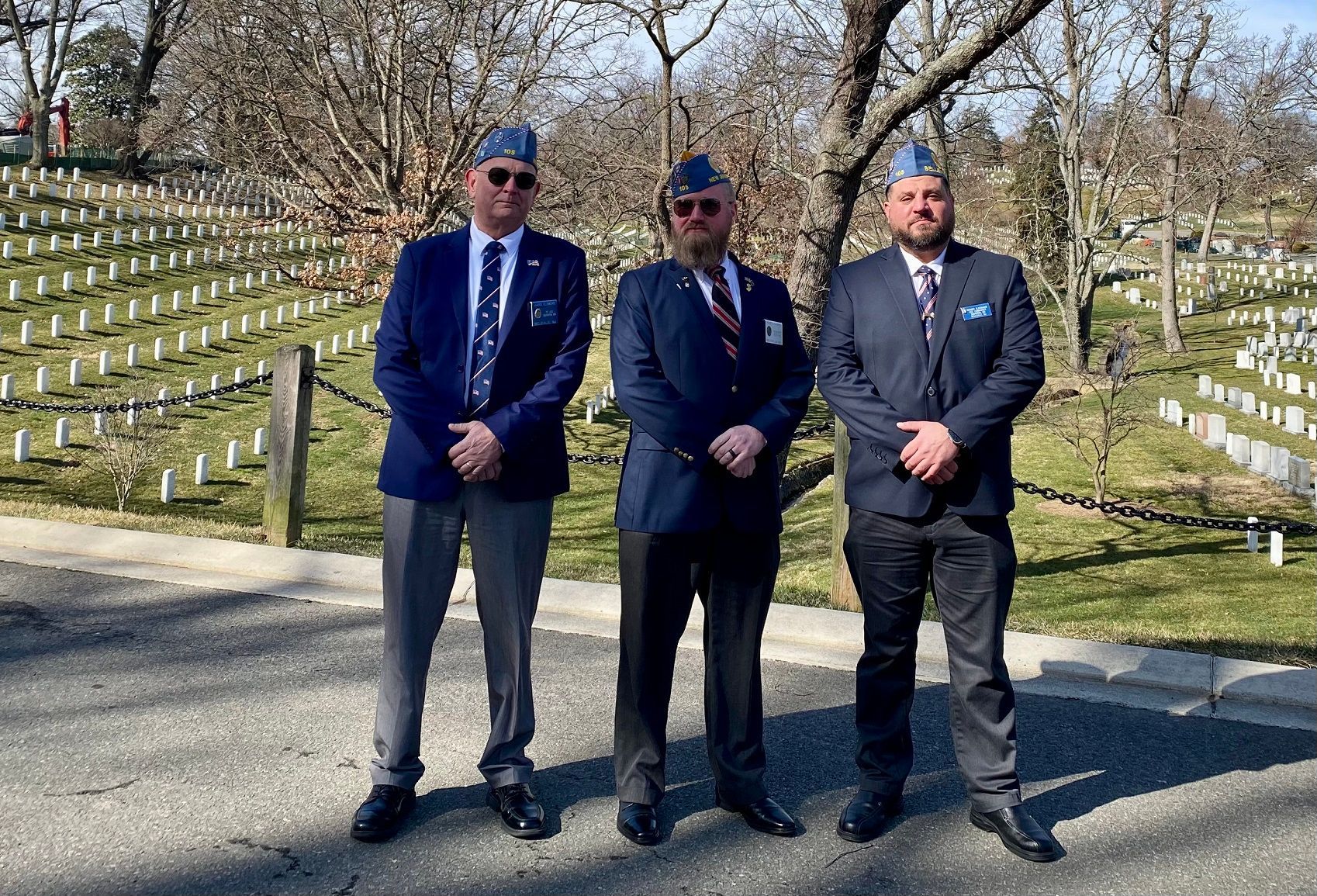 Three men in military-style jackets and caps stand in a cemetery. Rows of white headstones fill the background.