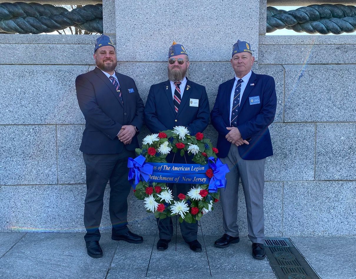 Three veterans in suits and hats stand beside a wreath in front of a stone wall.