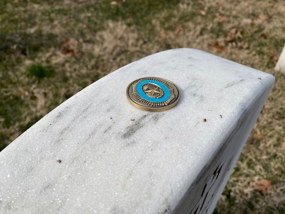 A coin with a teal ring rests on a white marble tombstone at a cemetery.