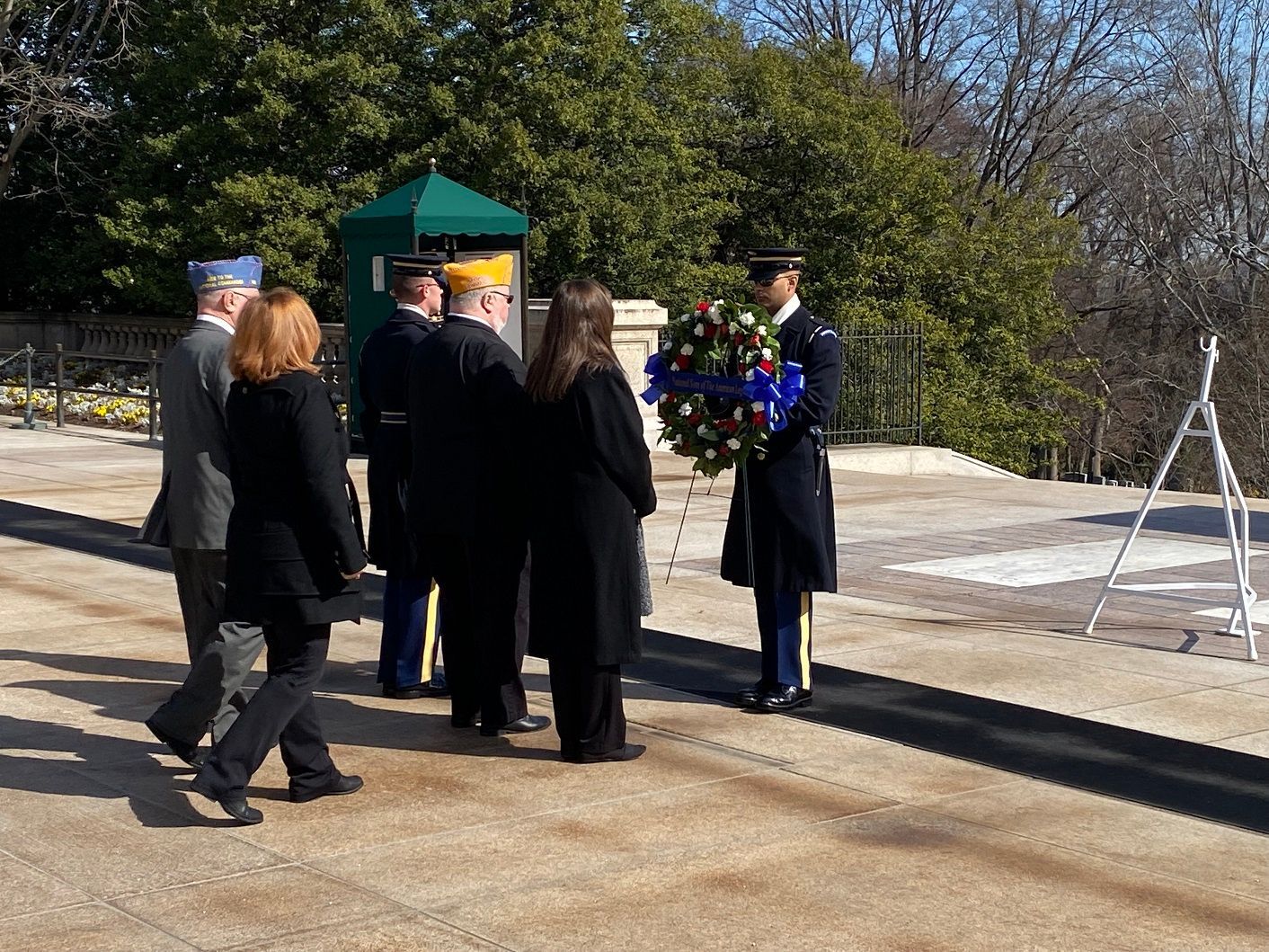 Group at memorial. Soldier holds wreath. Others watch. Sunny, outdoor setting.