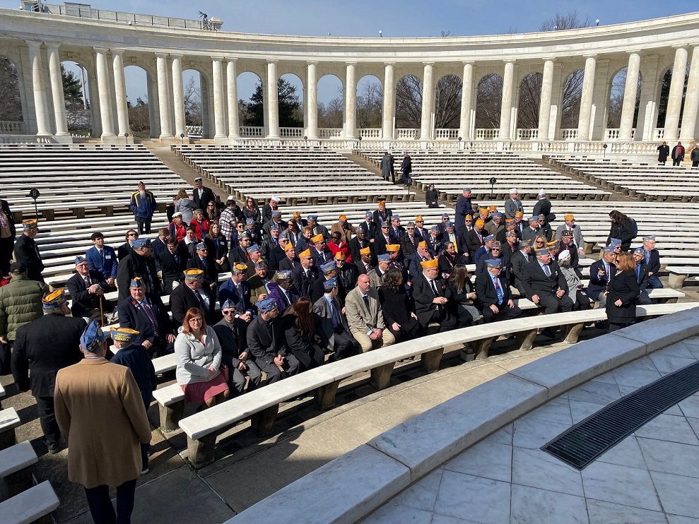 Group of people, many wearing caps, seated in an amphitheater with white columns and rows of seats.