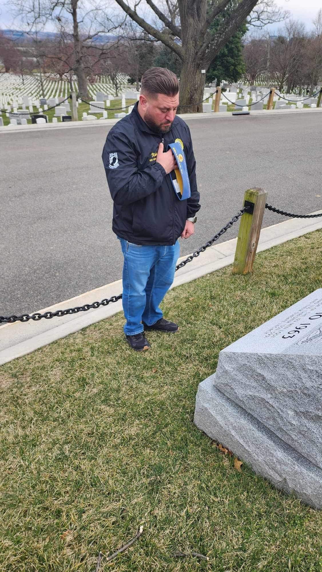 Man in navy jacket and jeans at a grave, hand on chest, Arlington National Cemetery.