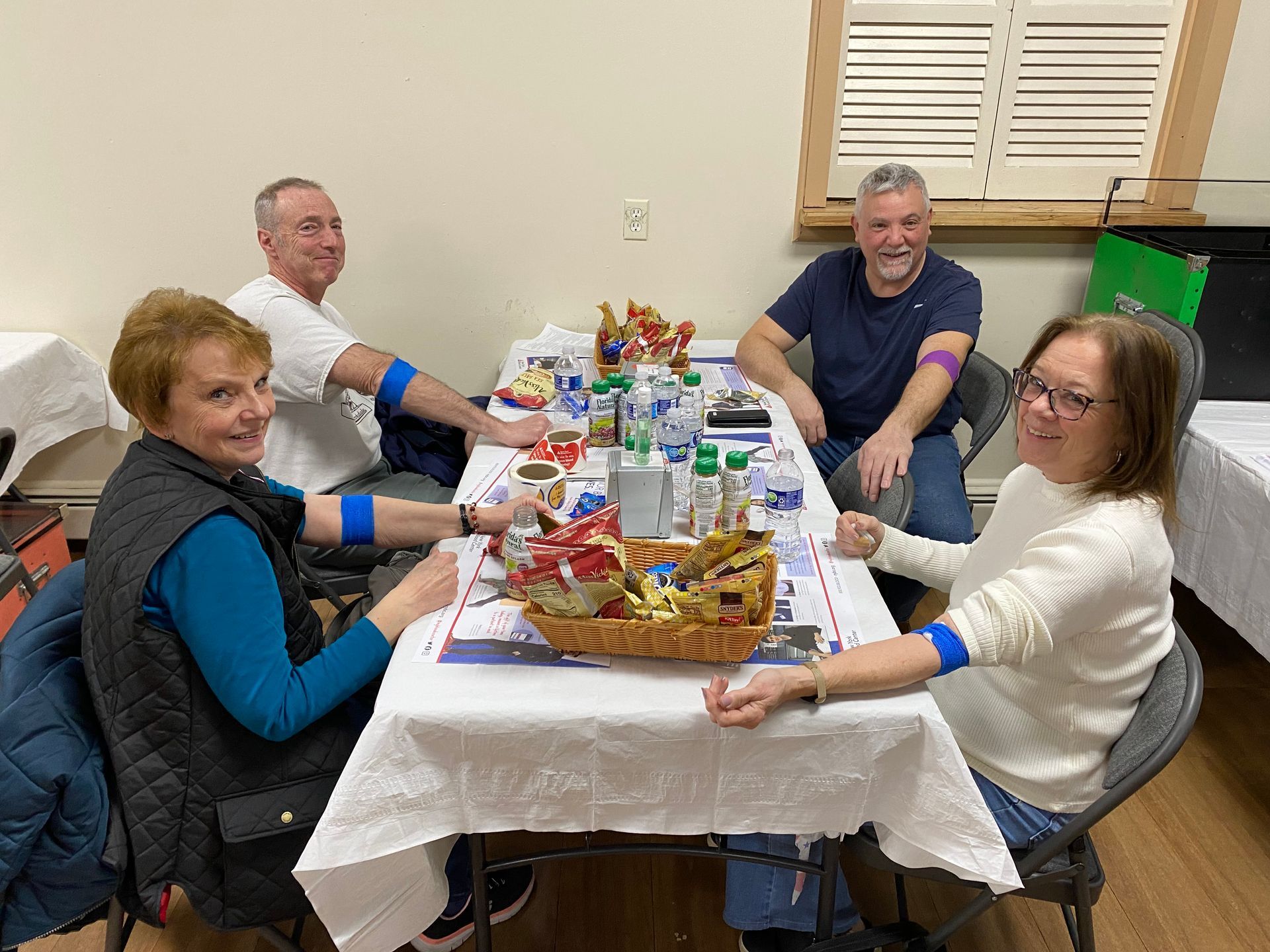 Four people sit at a table after donating blood. They have blue arm bands, snacks, and drinks.