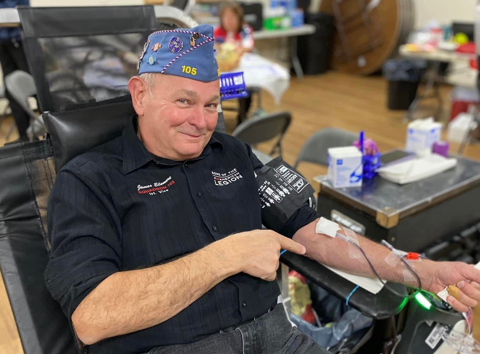 Man in black shirt donating blood, wearing a blue cap with 
