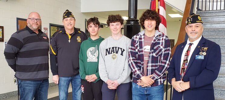 Group of men and boys pose, two wearing military hats. An American flag is in the background.