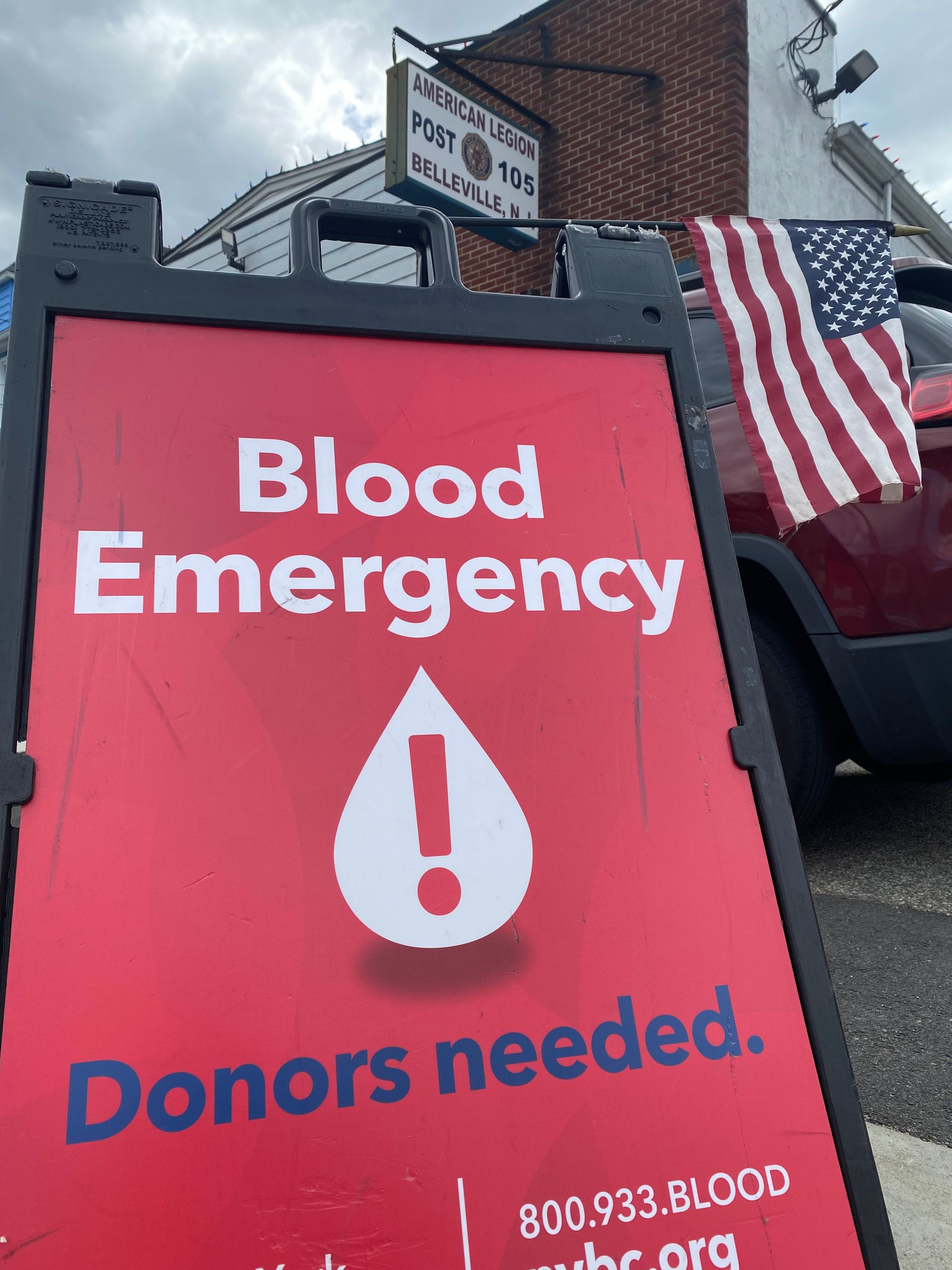 Sign: Blood emergency, donors needed; American flag, storefront backdrop.