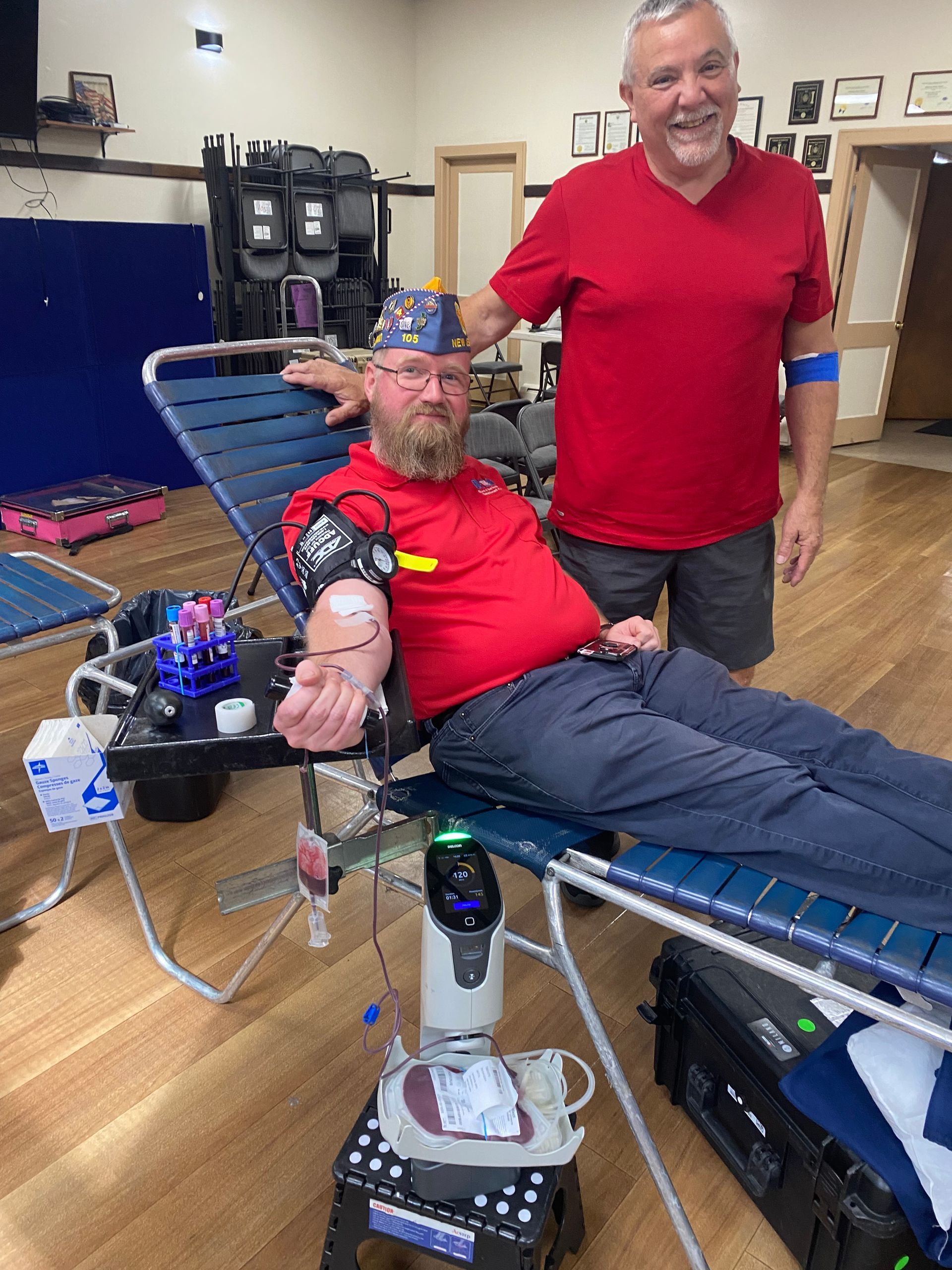Man donating blood, another man stands beside him. Both men wear red shirts.  Indoor setting with medical equipment.