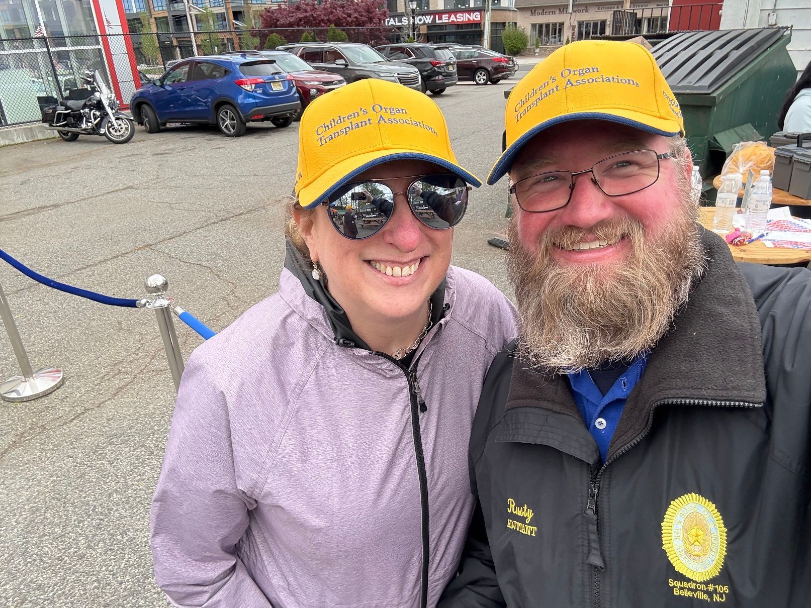 Two people wearing yellow hats smile at the camera. Outdoor setting, cars in the background.