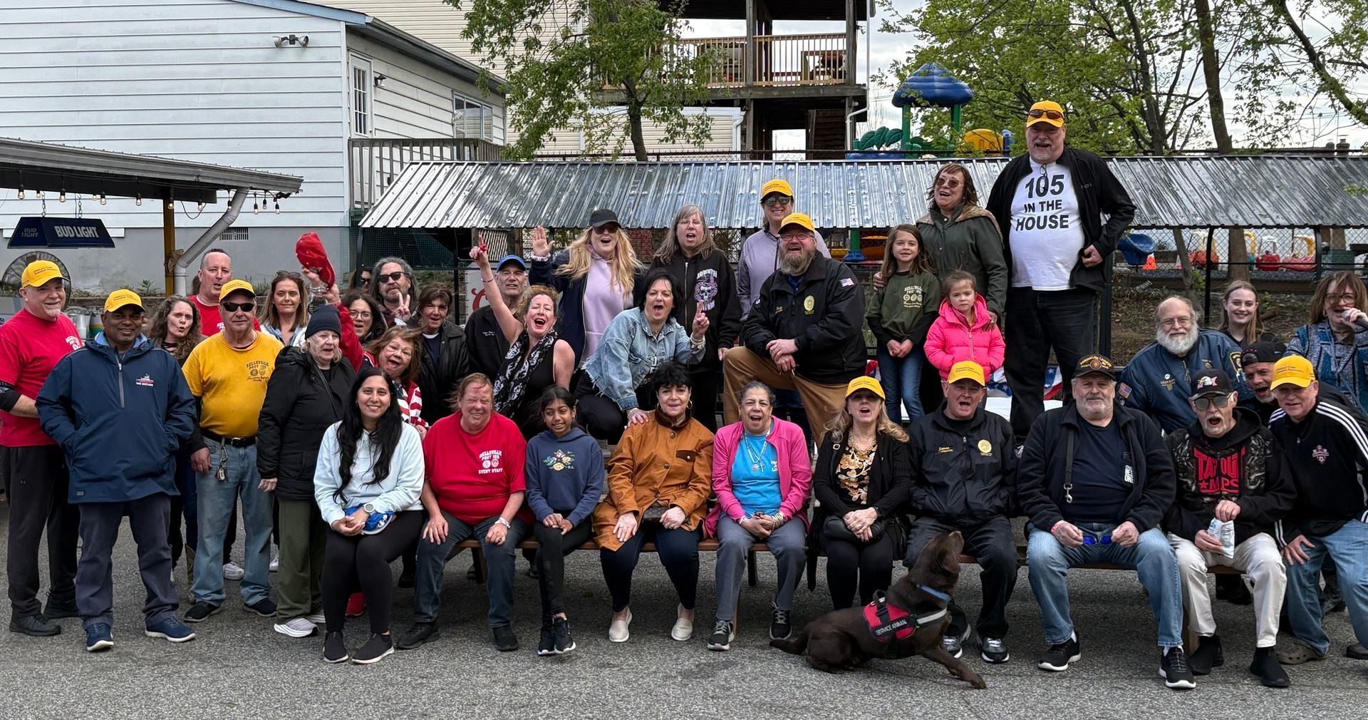 Group of people posing outdoors, many wearing yellow hats. Cloudy day, with buildings and a playground in the background.