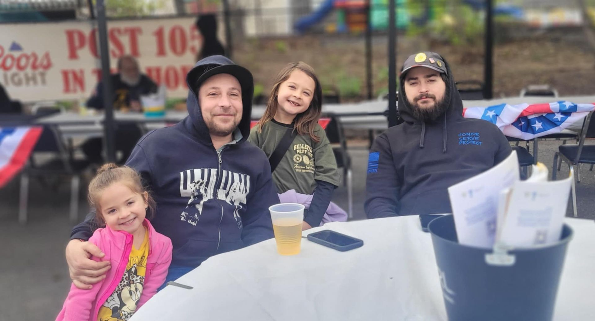 Two adults and two children at outdoor table; one adult is holding a child, smiles.