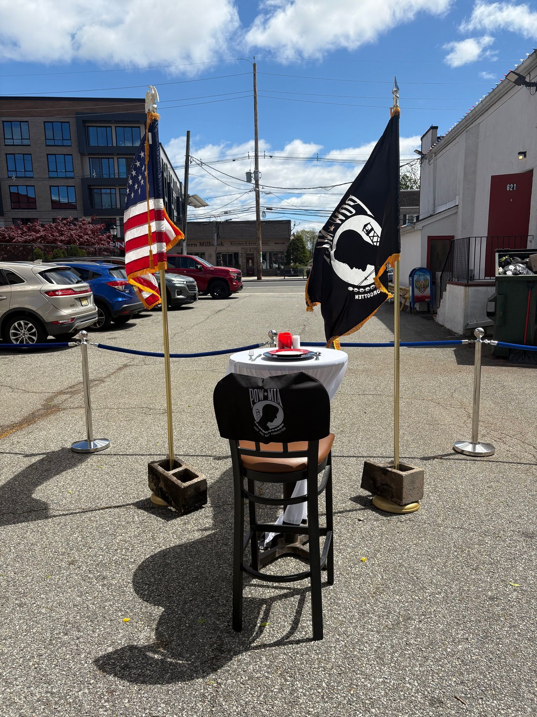An outdoor memorial with flags, a table, chair, and a shirt, set in a parking lot.