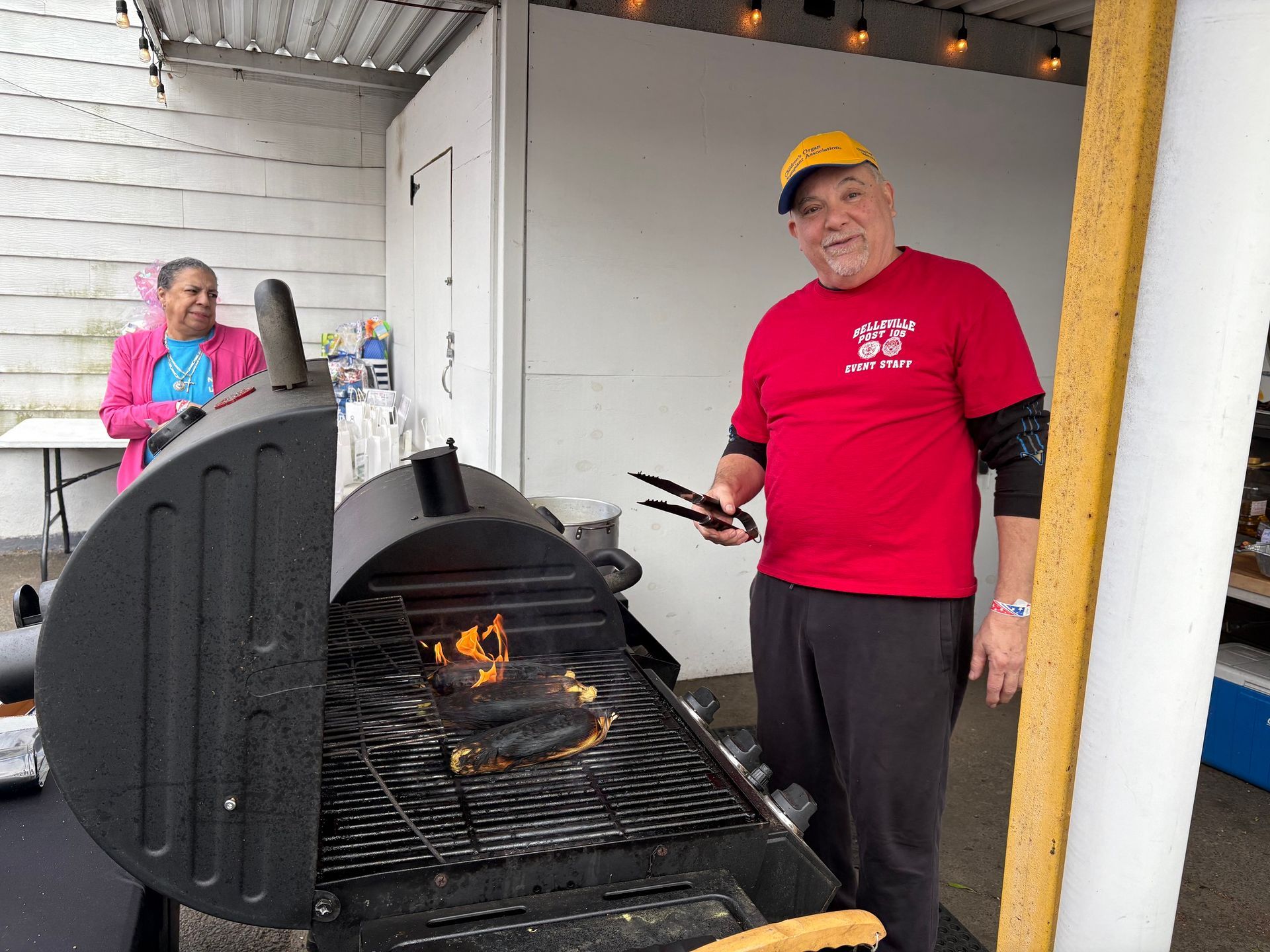 Man grilling food at a barbecue, wearing a red shirt and yellow cap. Another person is visible in the background.