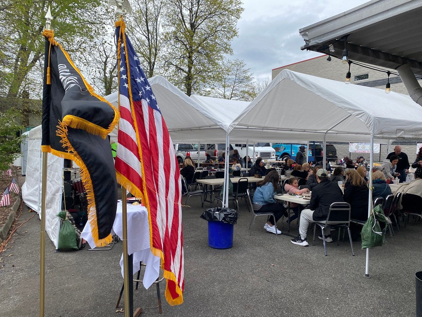 People seated at tables under tents, US and POW/MIA flags in the foreground.