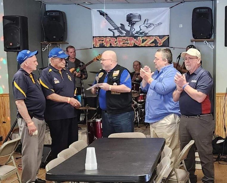 Men at a venue with a band, receiving a presentation. Some are clapping, one holds a paper.