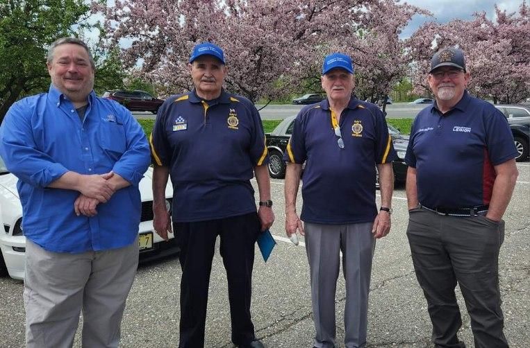 Four men in uniform standing in a parking lot. Two wear blue caps and polo shirts, two have button-down shirts. Cars and trees are in the background.