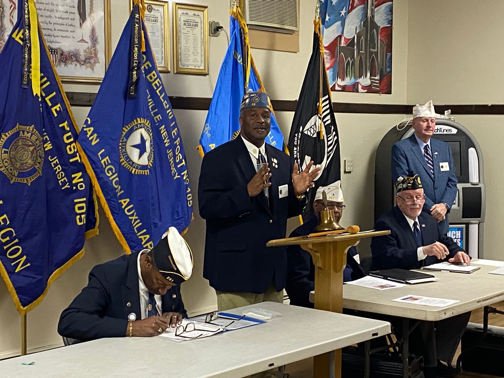 Men in military attire at a podium with flags, possibly at an American Legion meeting.