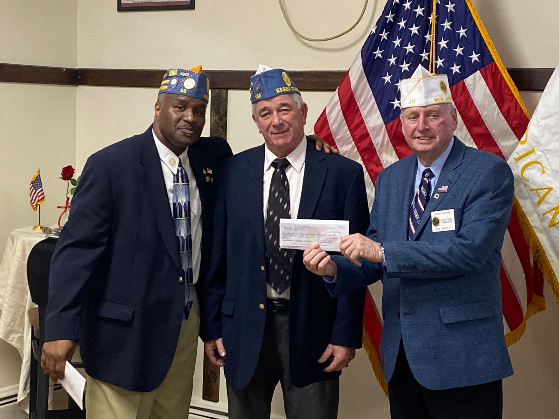 Three men in suits and hats stand in front of flags, one presenting a check.