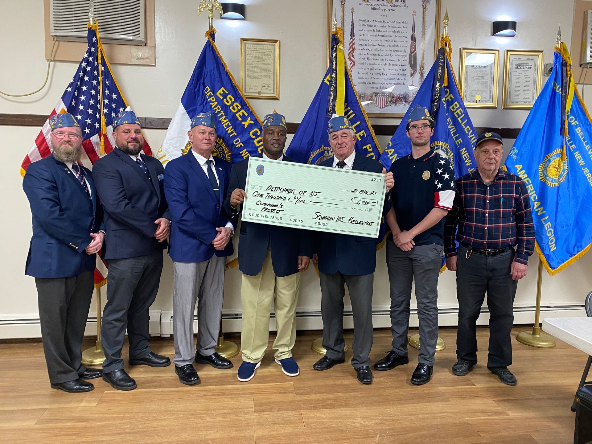 Group of men holding a large check, posing in front of flags and banners indoors.