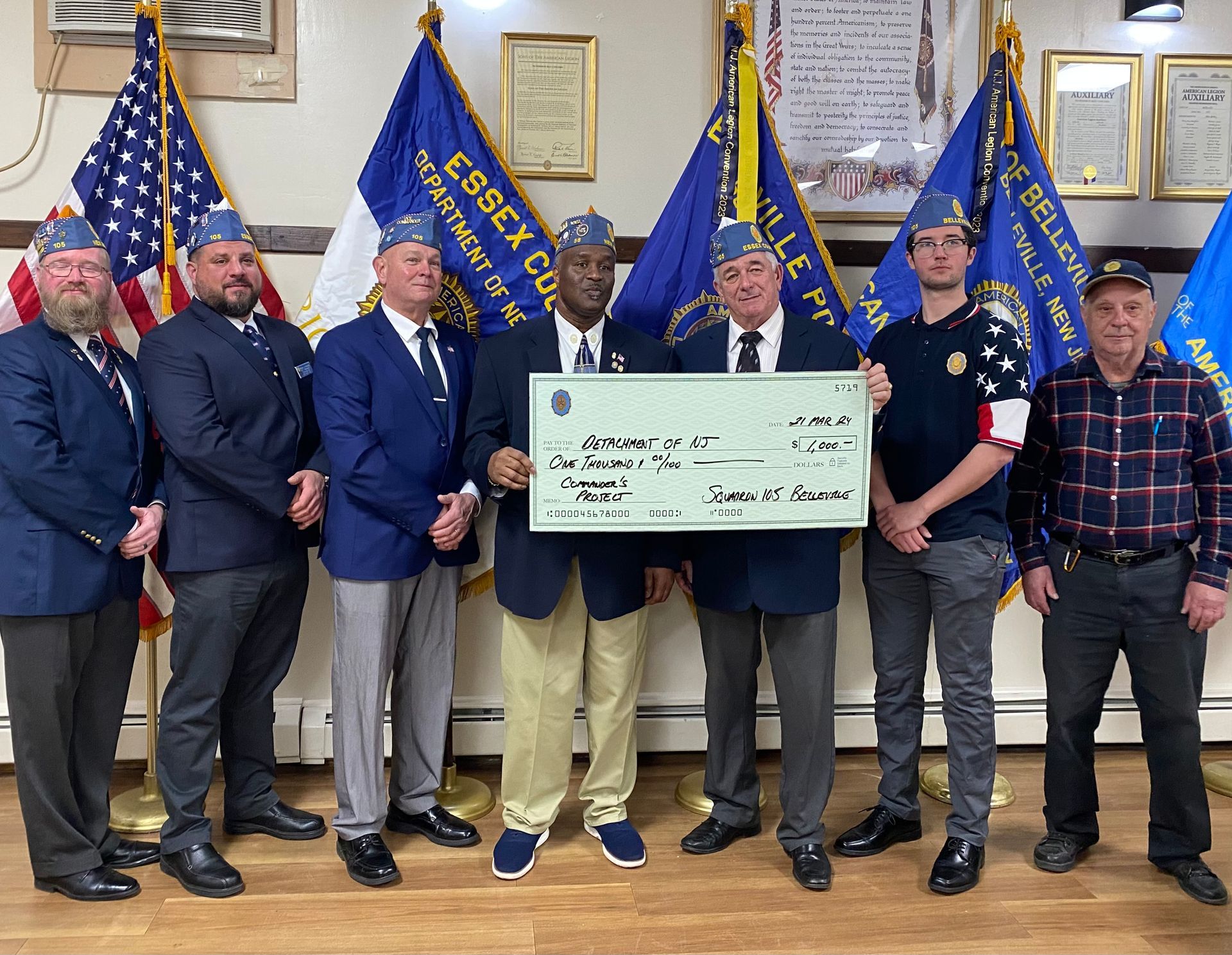 Group of people holding a large check, presumably a donation, in a veterans' hall with flags.