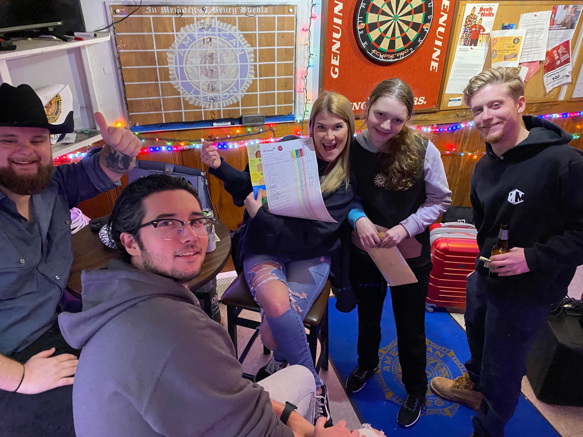 Group of five people celebrating, holding paperwork, in a bar with a dartboard and festive lights.