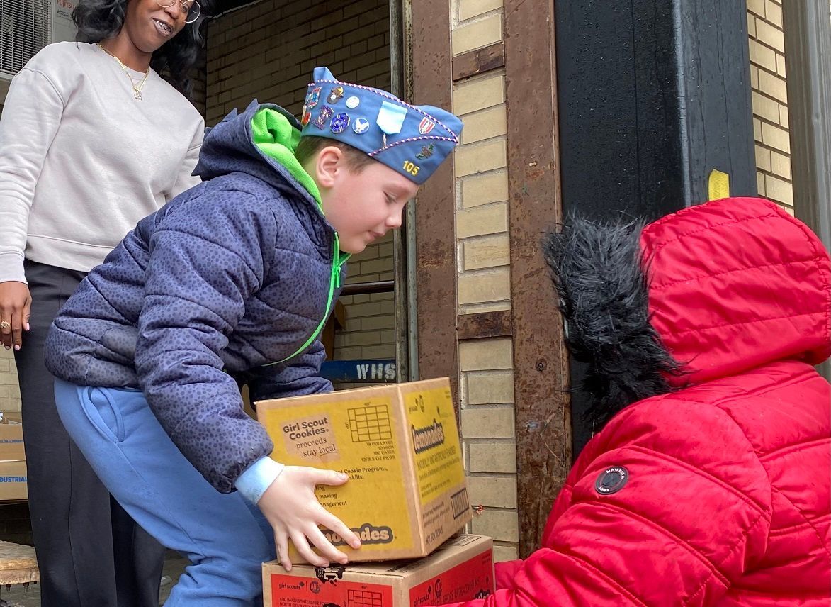 Boy in blue jacket and cap helps carry boxes outside. Person in red jacket assists. Woman watches.