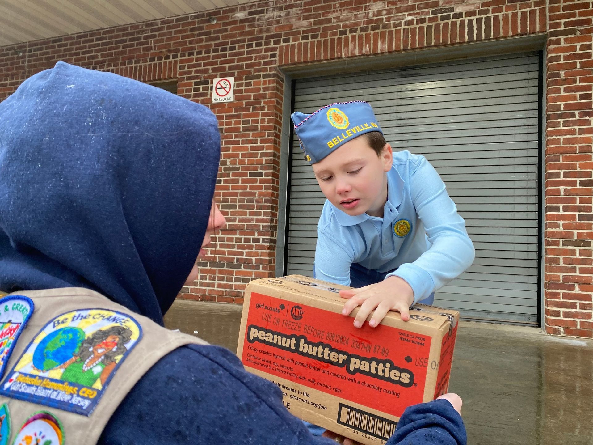 Two boys exchange a box of peanut butter patties outside a brick building. One wears a scout vest and the other a scout hat.