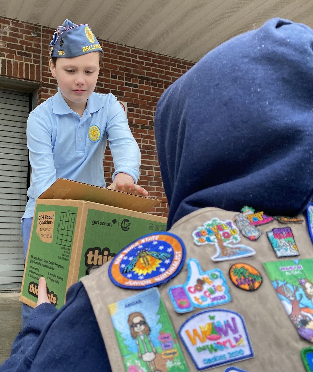 Boy in uniform opens a box held by a person wearing a scout vest with badges outside a brick building.
