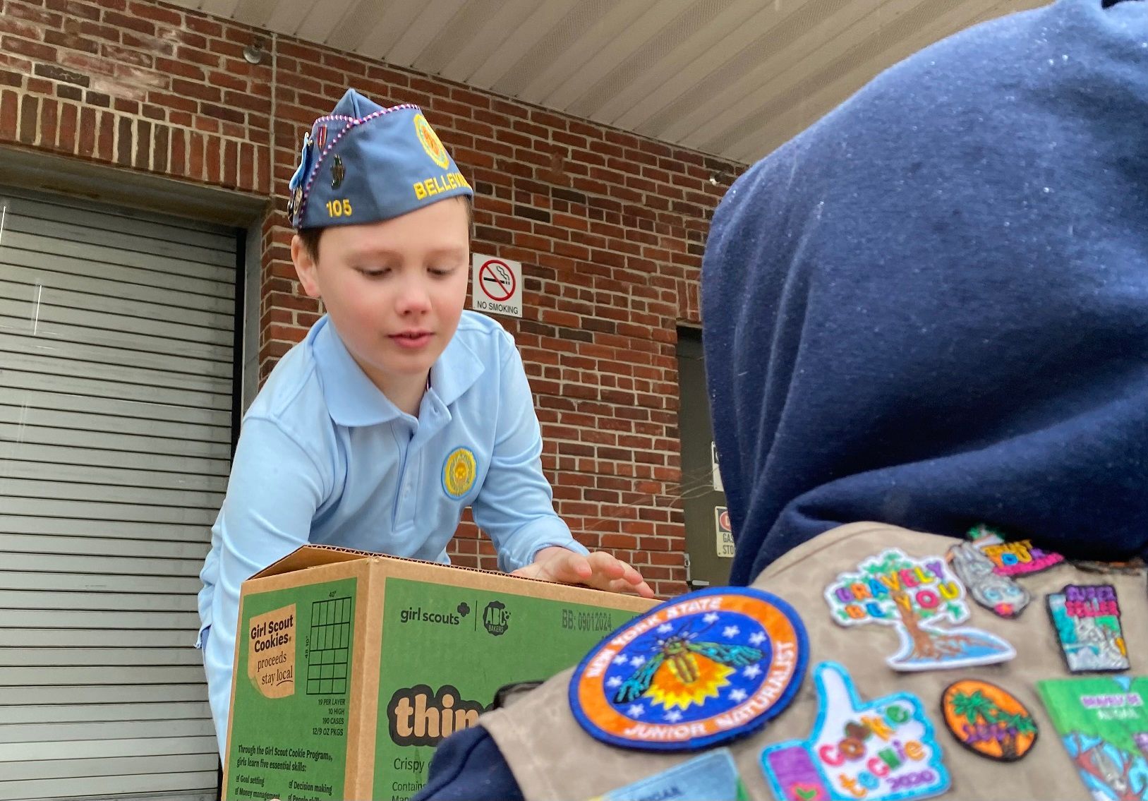 Boy scout in blue uniform handling a box, standing next to another person with scouting patches, brick building in background.