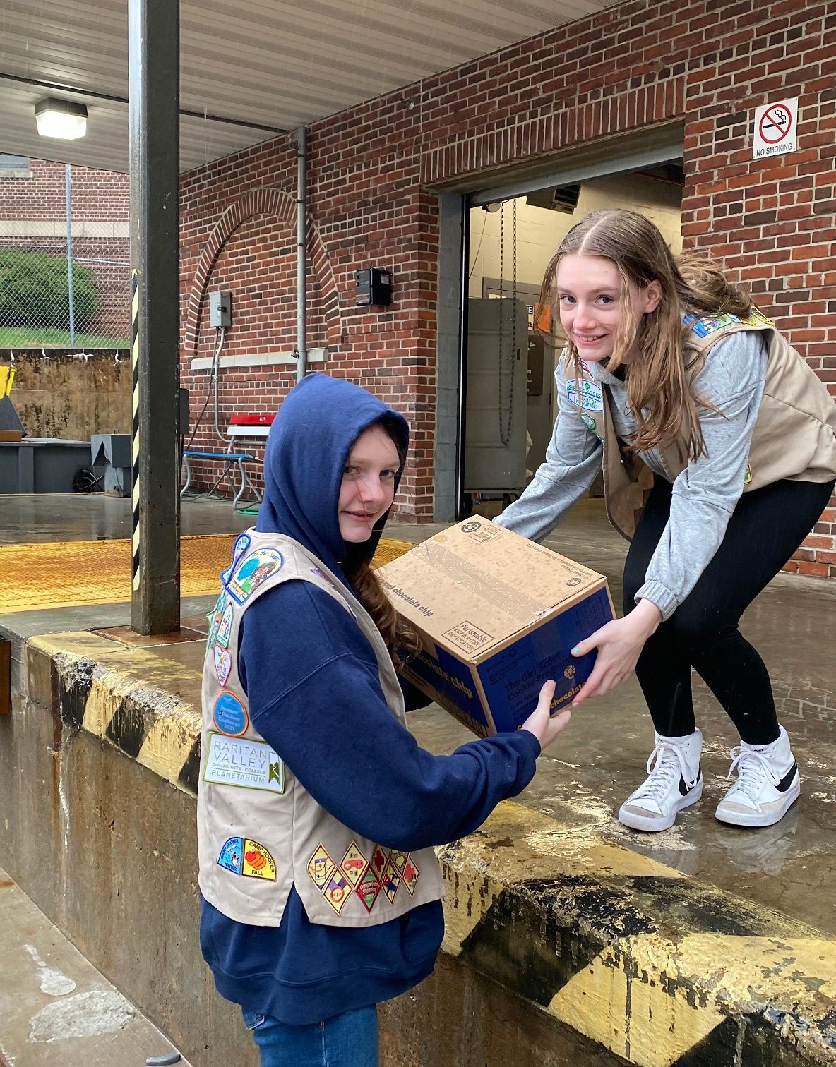 Two Girl Scouts in vests pass a cardboard box at a loading dock.