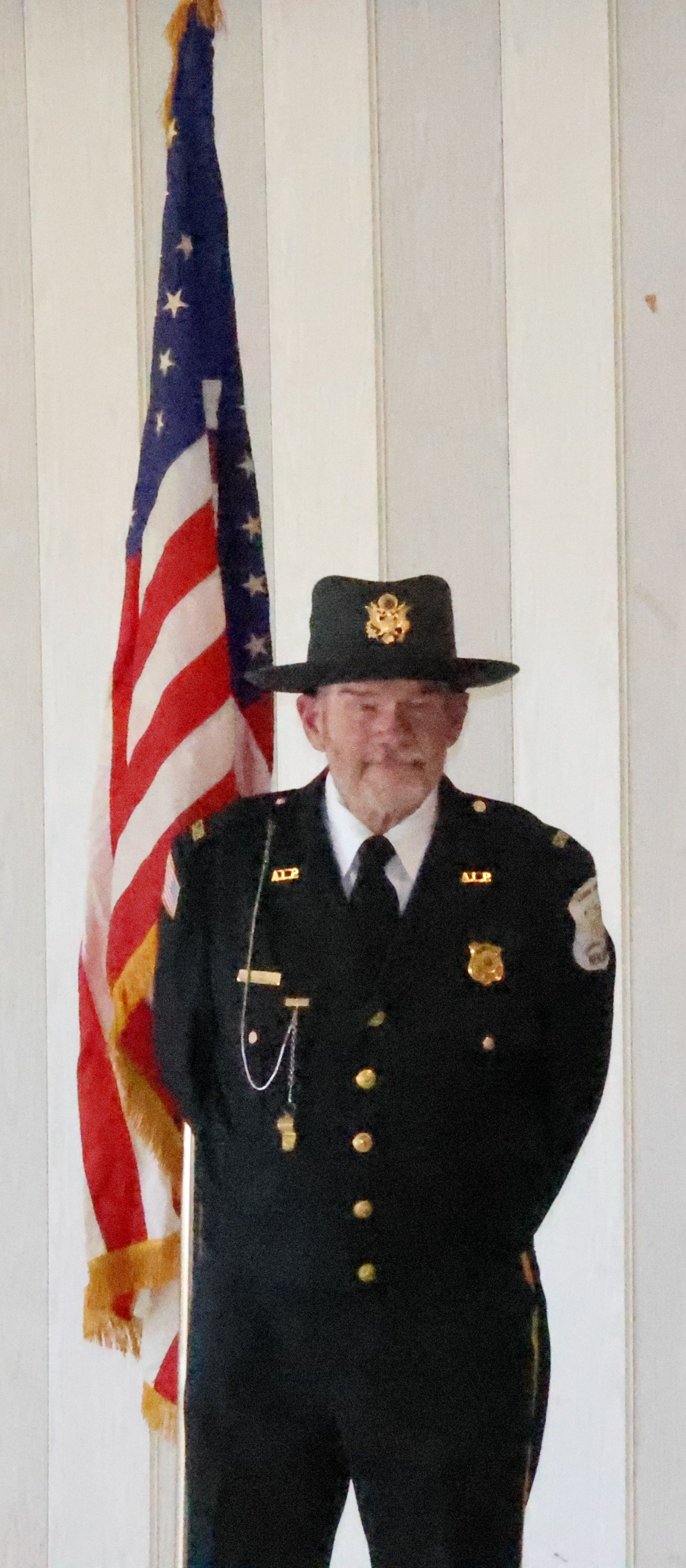 Man in dark uniform with a hat stands in front of an American flag. He has his hands behind his back.