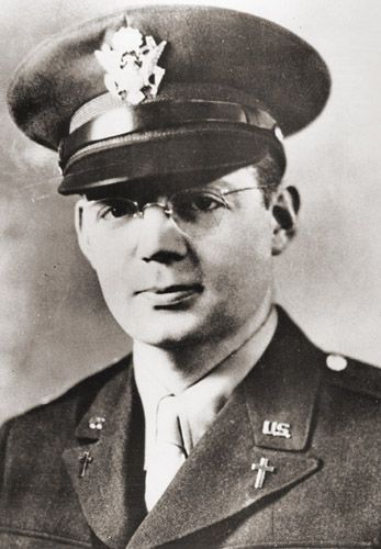 Man in military uniform, wearing glasses and a hat with a pin. He has a serious expression.