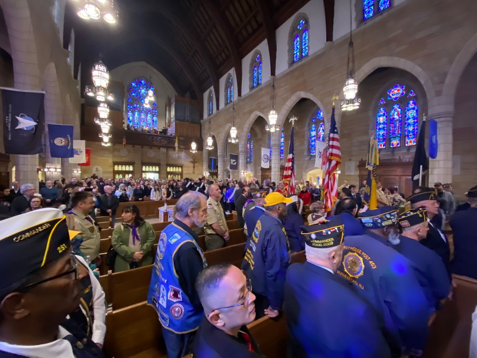 Inside a church, veterans and others attend a ceremony with flags and stained-glass windows.