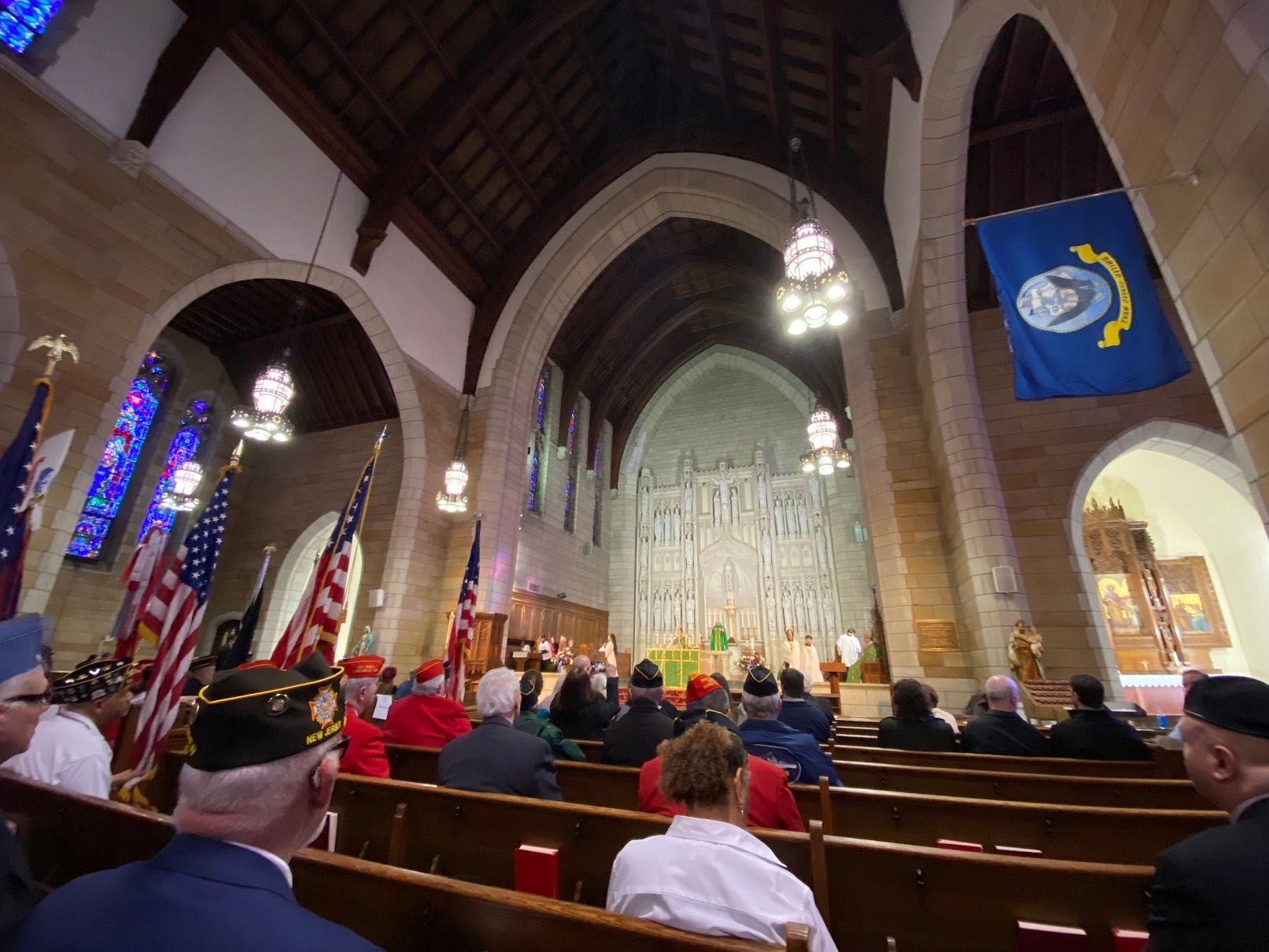 Interior church scene with people seated, flags, altar, and arched ceiling.