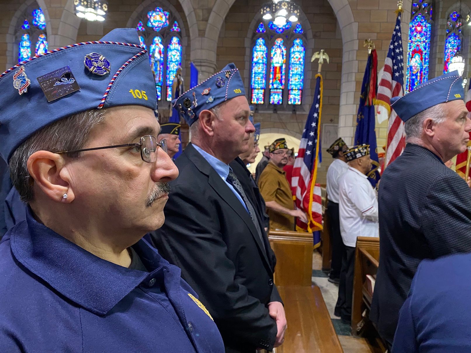 Veterans in blue caps stand inside a building with stained-glass windows and flags.