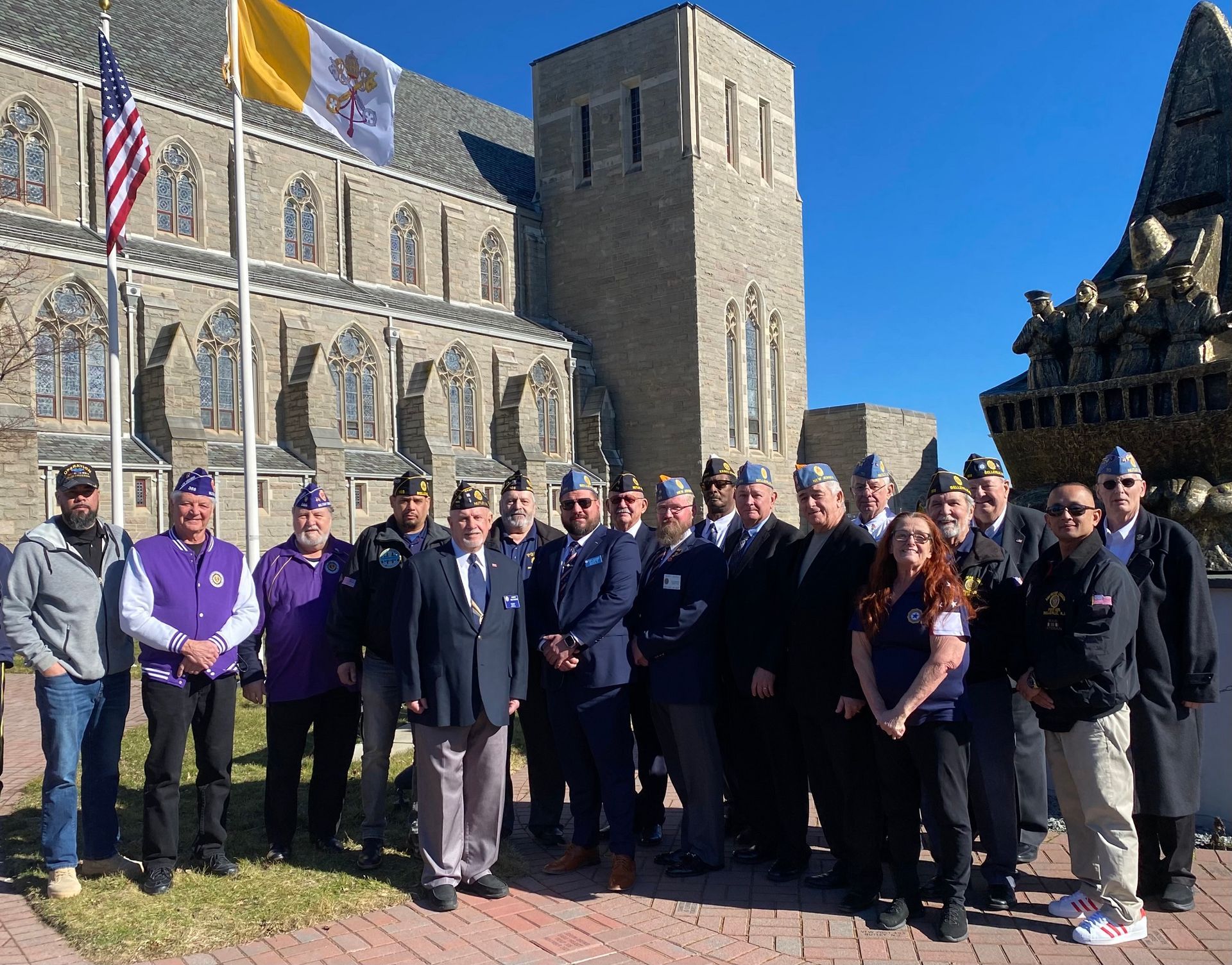Group of veterans in uniform pose in front of a stone church and a memorial sculpture under a clear sky.