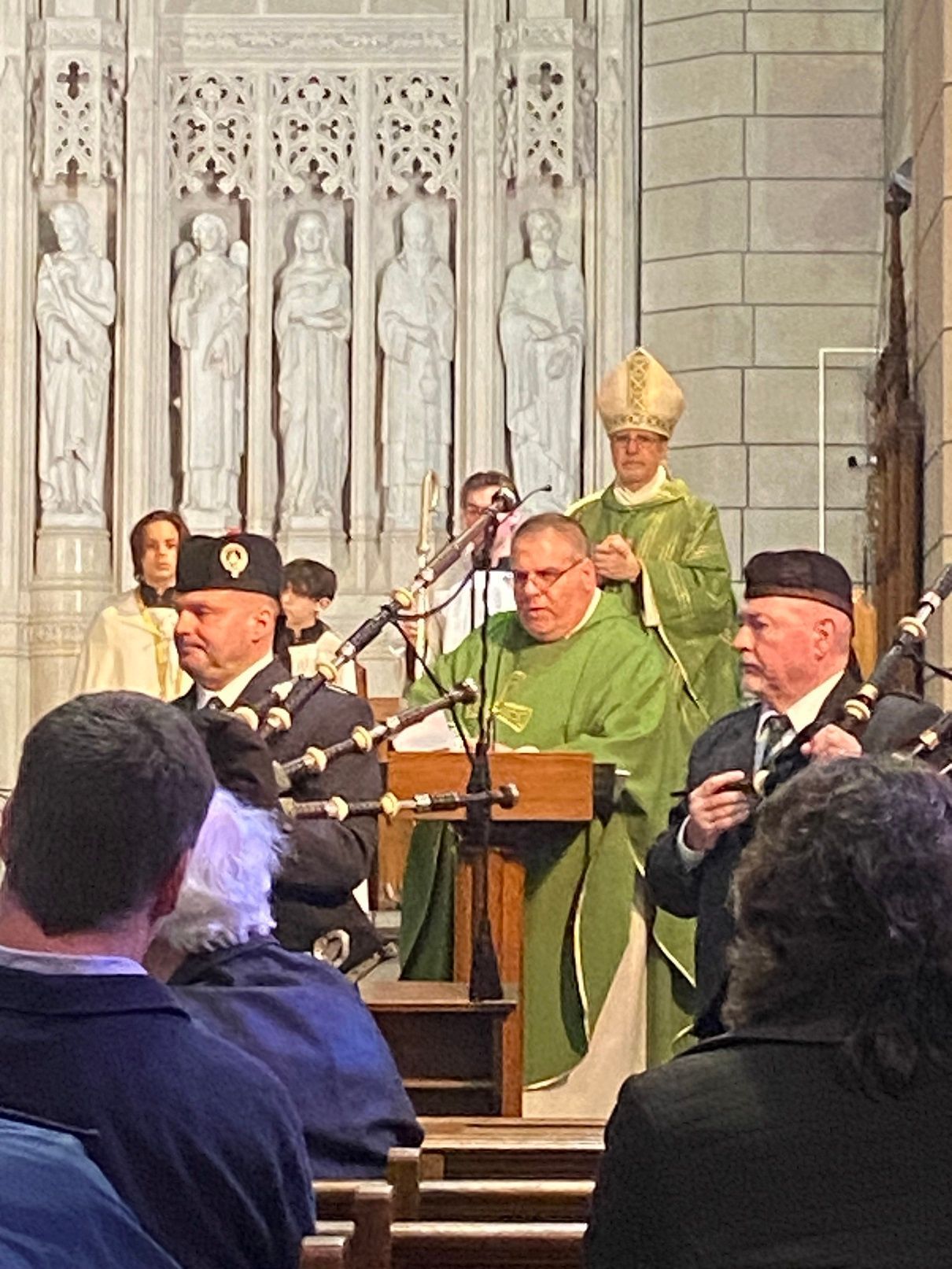 Clergy leading a service in a church, flanked by bagpipers, with attendees in pews.