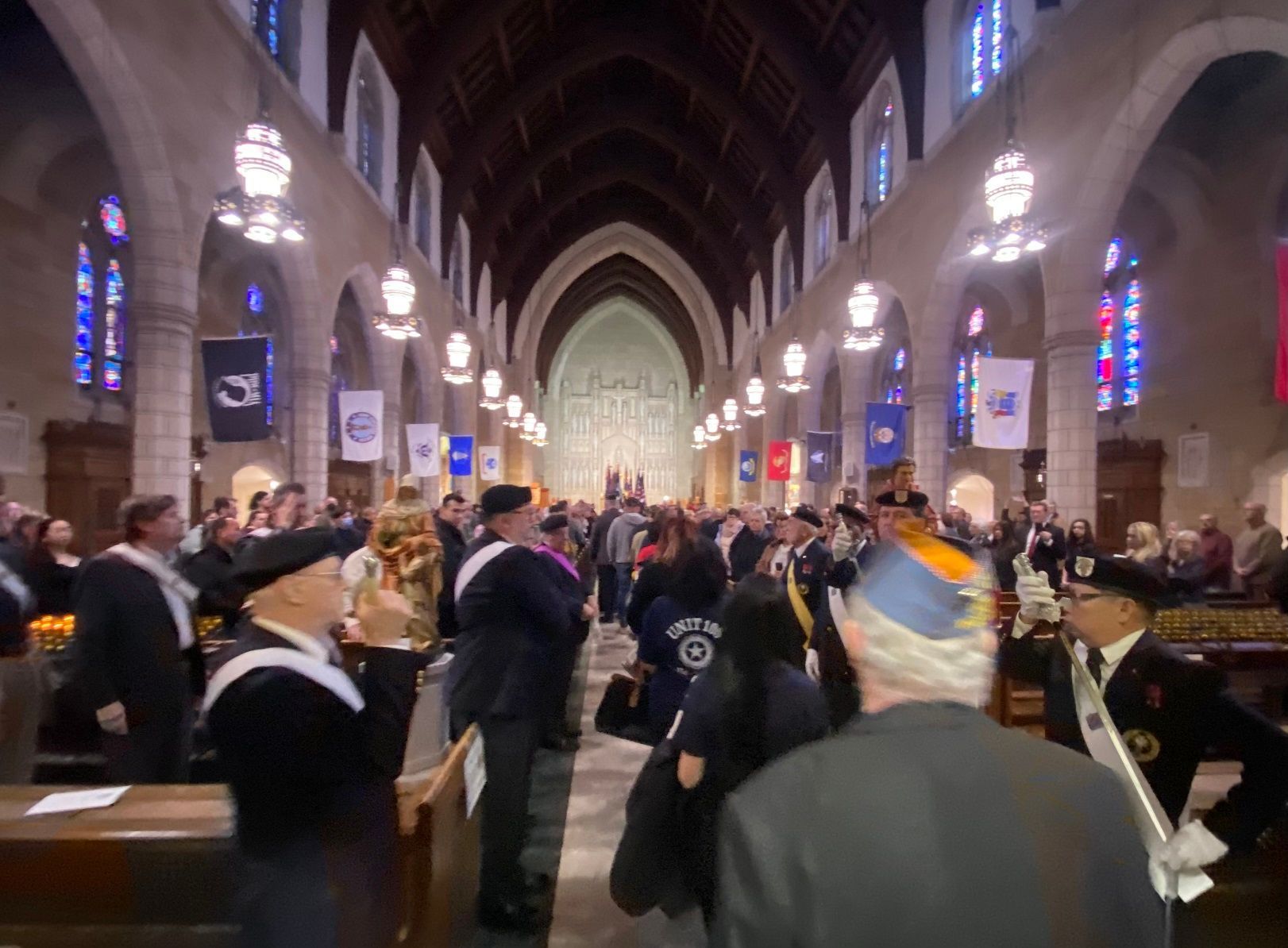 Interior of a church with people standing. Various banners hang from the walls. Some wear formal attire with hats.