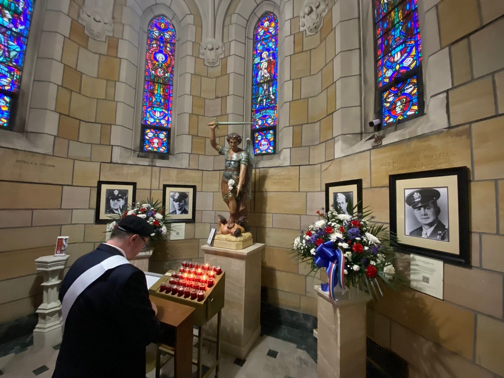 A person in uniform reads at a candlelit altar, a statue, stained glass windows, and framed photos are present.
