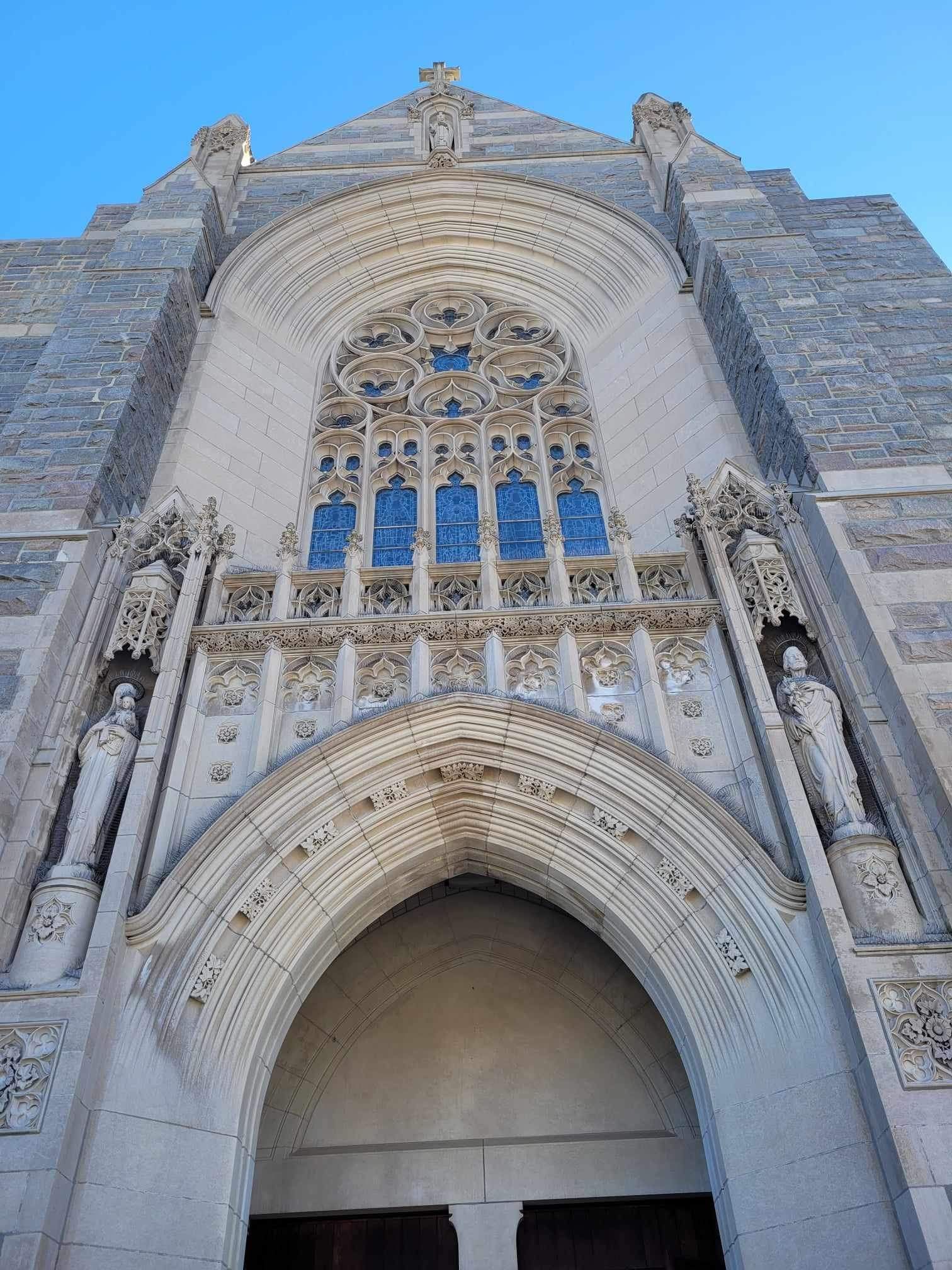 Stone church entrance with arched doorway and large stained-glass window under a clear blue sky.
