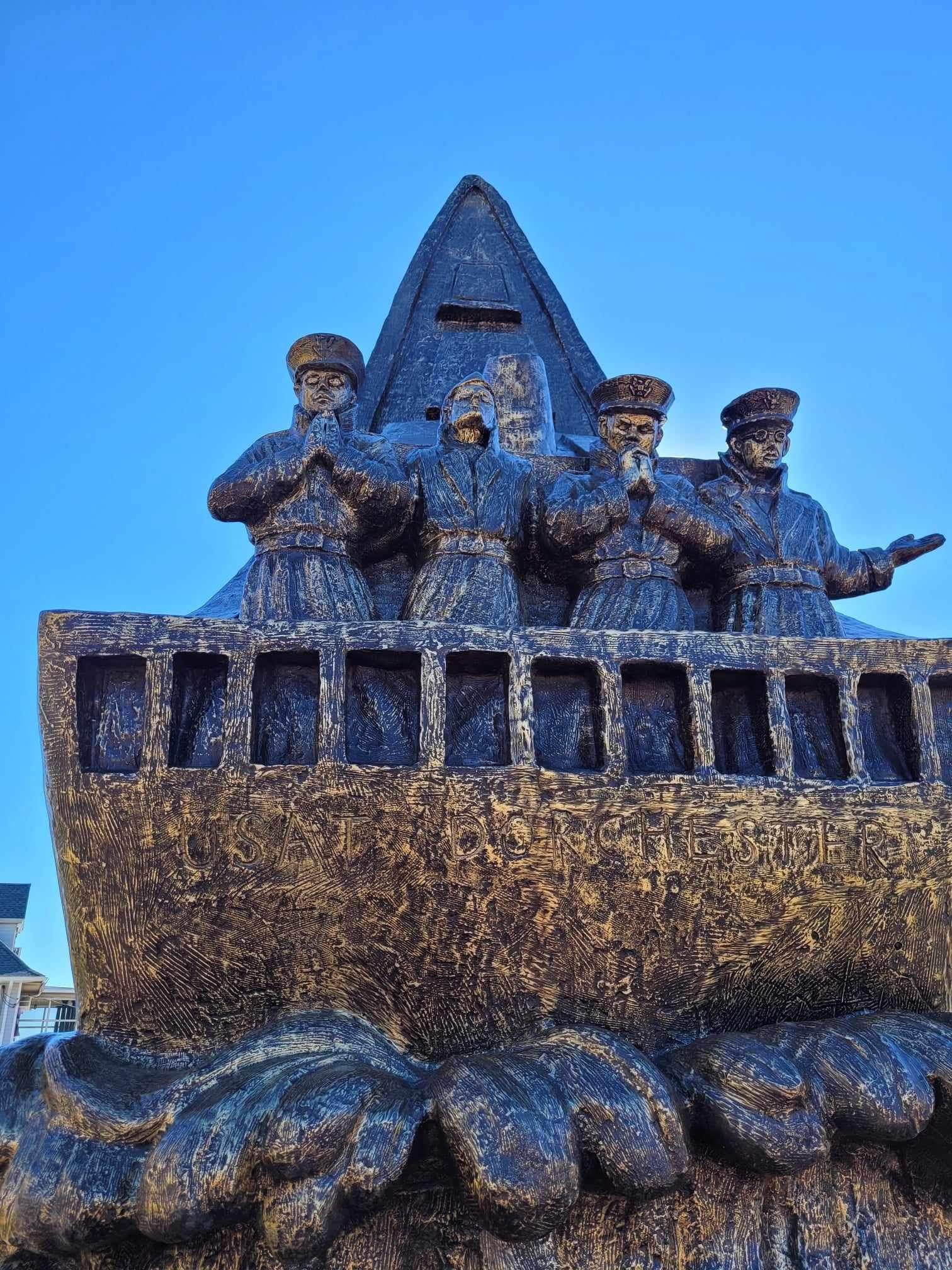 Monument of four figures on a ship, facing forward against a blue sky. Waves at the base.