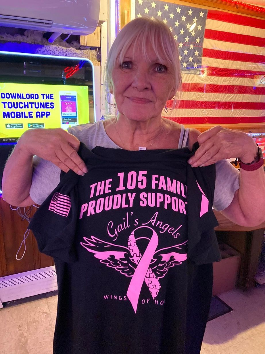 Woman holding a black shirt with pink text and ribbon, American flag in background.