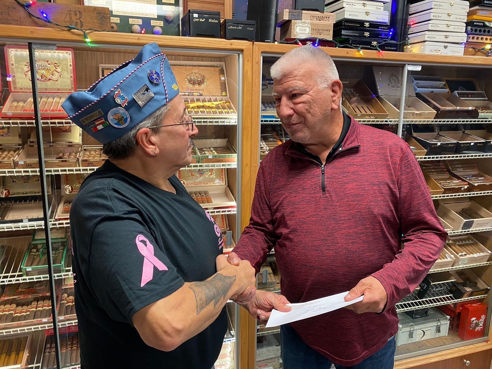 Two men shaking hands in a cigar shop. One wears a blue cap and a breast cancer awareness shirt. The other holds a paper.