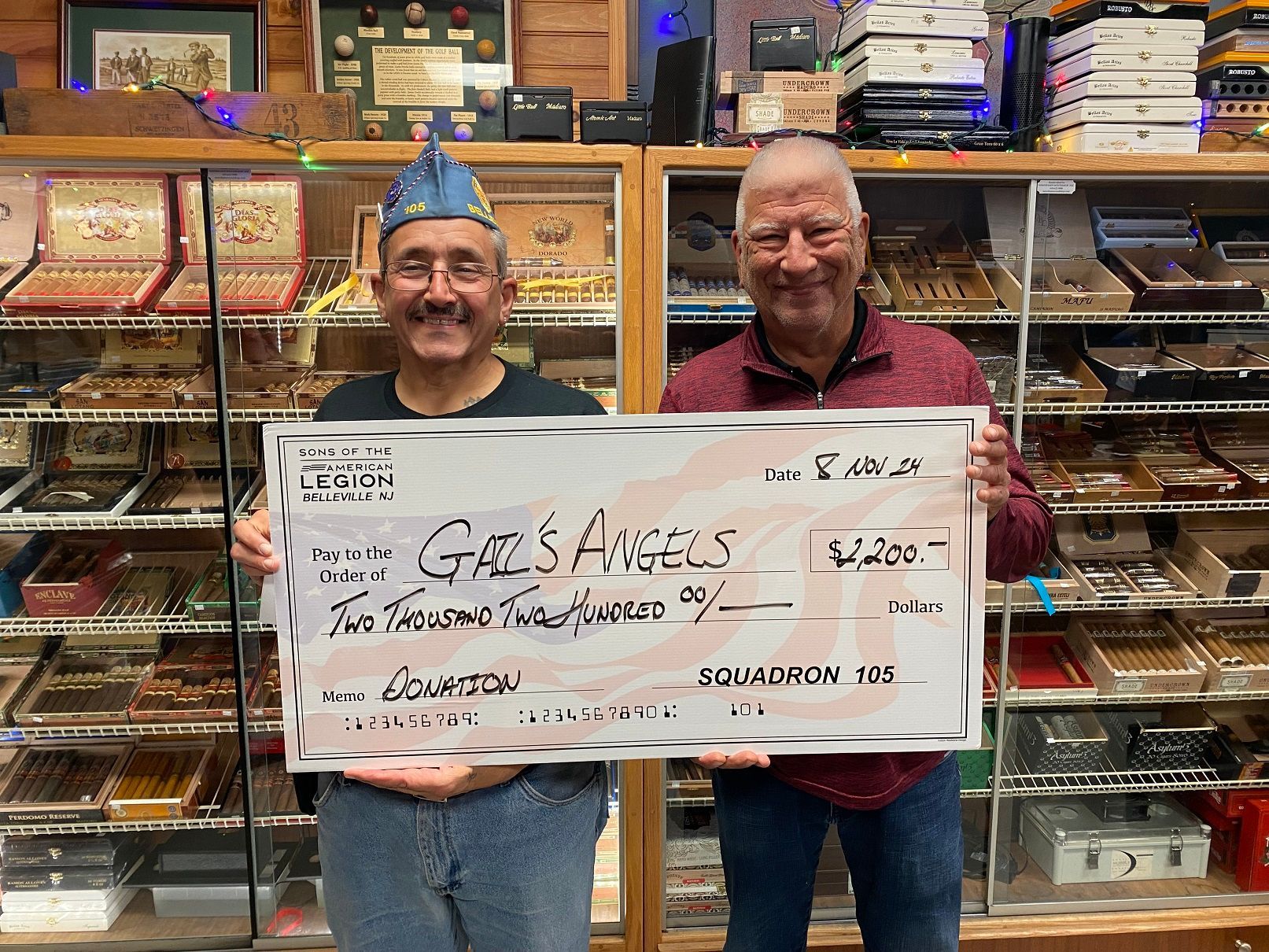 Two men hold a large check for $1200 in a cigar shop.