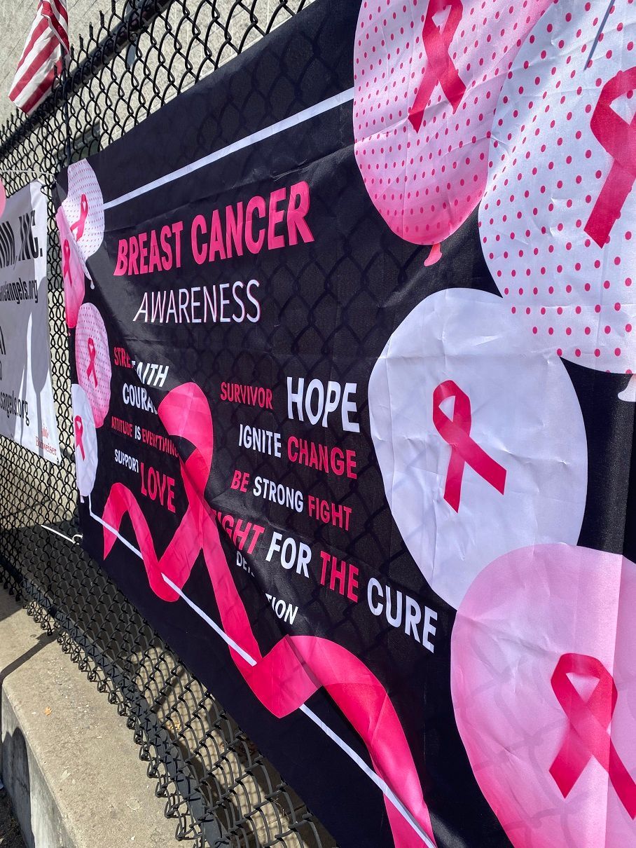 Black banner with pink breast cancer awareness ribbon symbols and text against a chain-link fence.