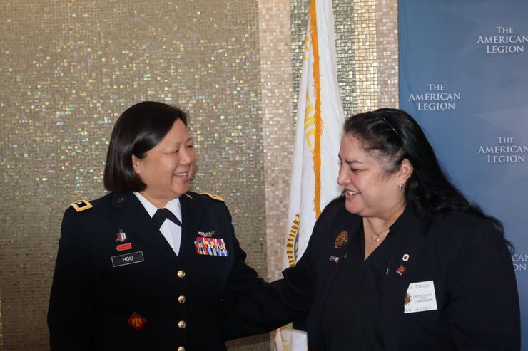 Woman in military uniform and woman in civilian attire smiling, side by side near an American Legion banner.