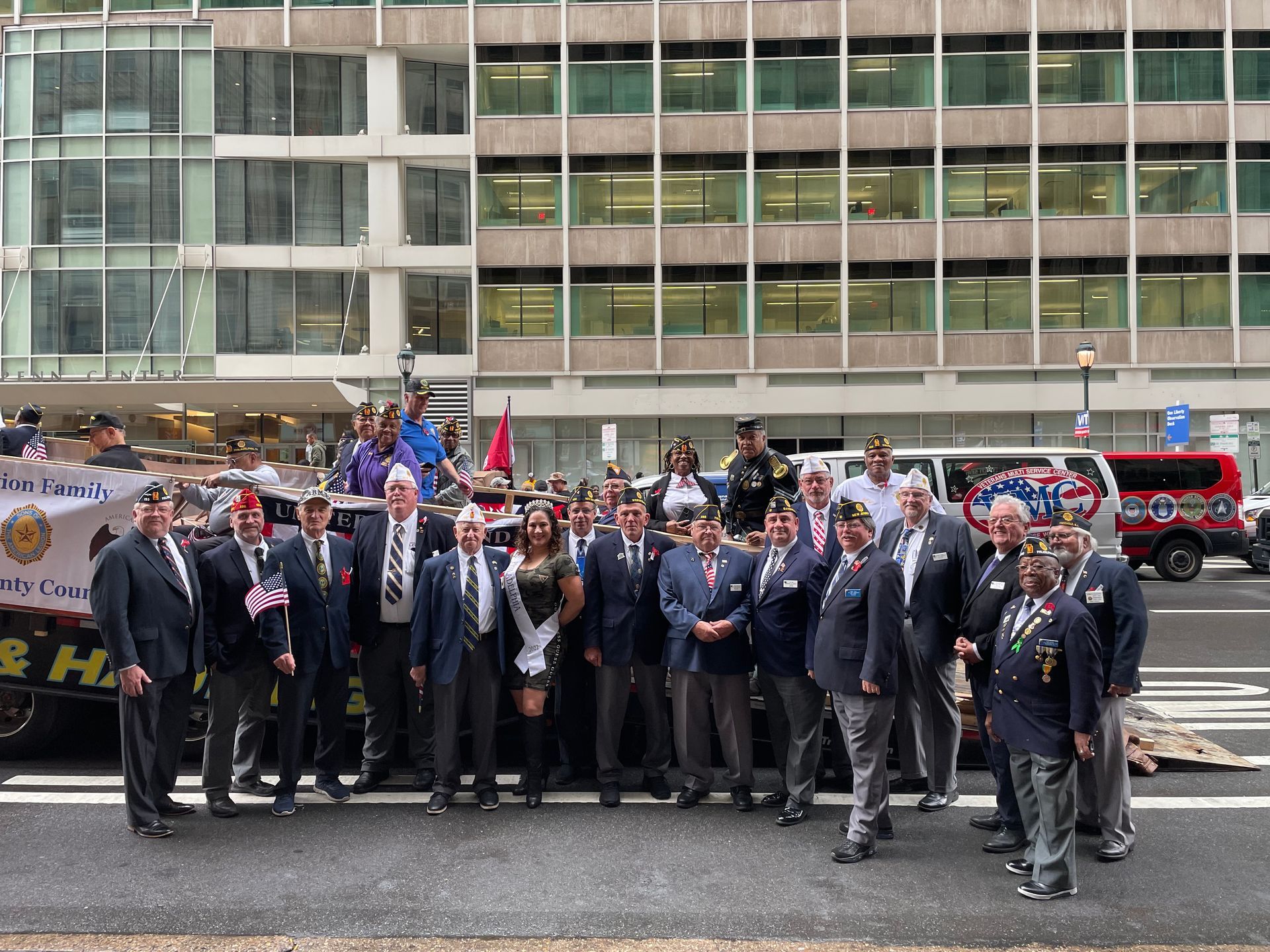 Group of veterans in suits posing in front of a building, some wearing hats and ribbons.