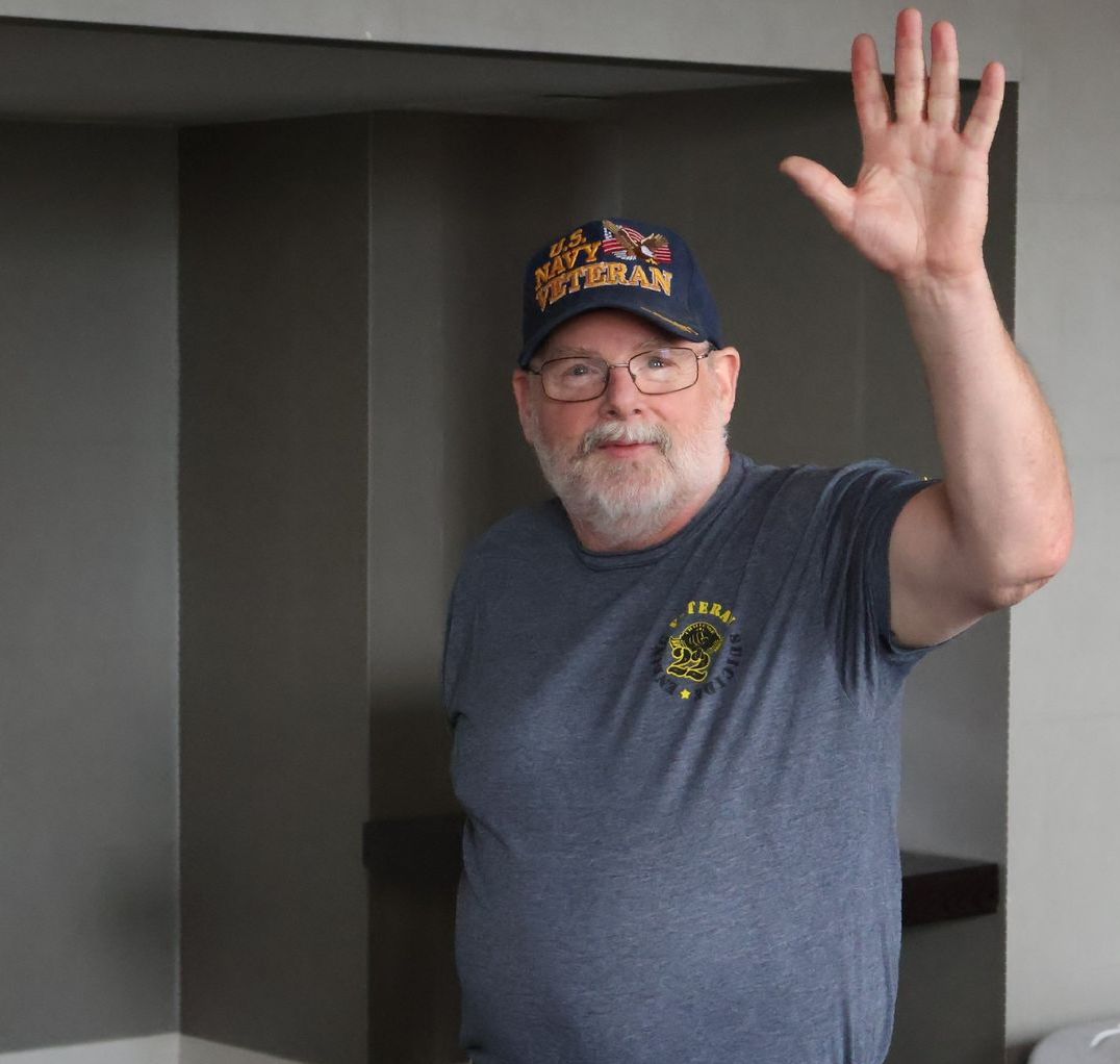 Man in blue Veteran hat waves. Gray wall and doorway in background.