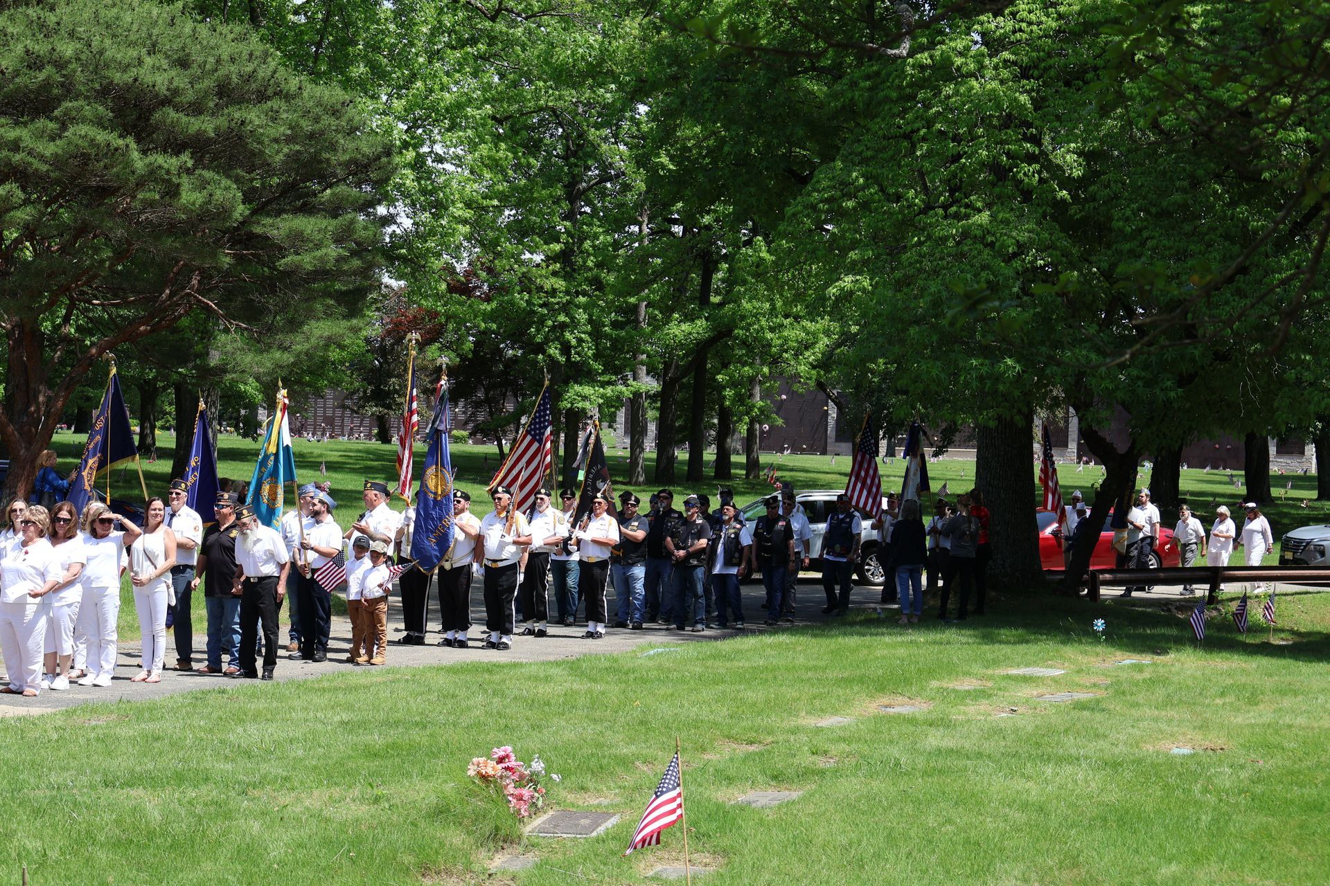 Crowd gathered in a park for a ceremony, with flags and a marching band under trees.