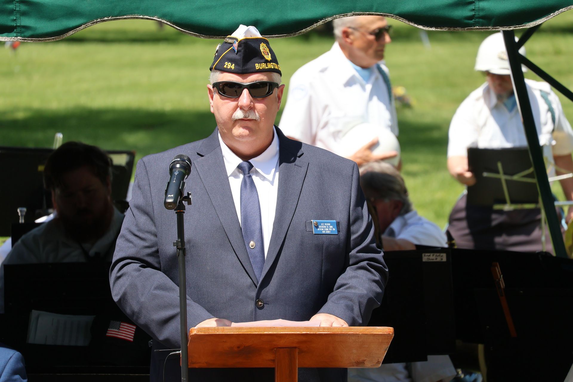 Uniformed man speaking at a podium outdoors, with musicians seated behind him.